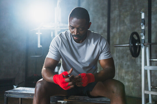 Fitness workout in gym. African American man fighter preparing for fight wrapping hands with red boxing wraps sports protective bandages in gym. Strong man ready for fight boxing sparring training