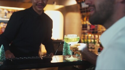 Special drink in Midori Sour cocktail with caramelized citrus slice and cinnamon power served stylish saucer glass by bartender serving to smart guest at luxurious bar christmas tree theme. Vinosity.