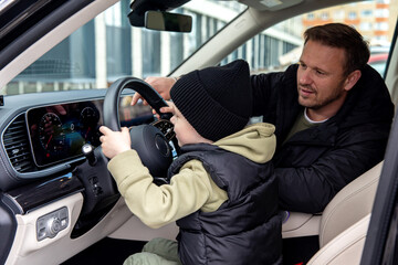 Happy little boy sitting behind the wheel of a car in the driver's seat while his dad is next to...