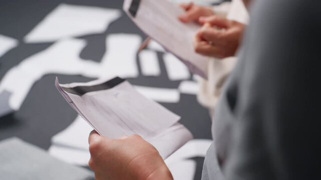 Closeup view of stylists demonstrating cloth designs drawn on white paper sheets during creative planning session, holding pattern sketches while fabric samples are visible in workspace indoors