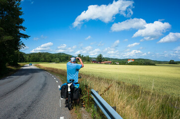 A bicycle traveler stops by the roadside in V&auml;stra G&ouml;taland County, Sweden, taking photo of the scenic countryside view. Golden fields and a bright summer sky create a peaceful rural landscape