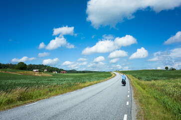 A scenic countryside road winds through open fields in Västra Götaland County, Sweden, under a bright blue sky. A touring cyclist rides in the distance, enjoying the peaceful summer landscape