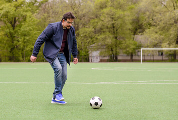 Obraz premium Middle-aged man in casual clothes playing soccer on green footbal field, ready to kick ball. Trees and goal posts in background. Active outdoor lifestyle and weekend recreation for adults. Copy space