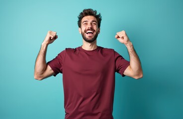 Fototapeta premium Young caucasian man on blue background cheers with raised fists celebrating victory. Happy guy is excited and smiling with arms up feeling great joy and success. Studio shot.