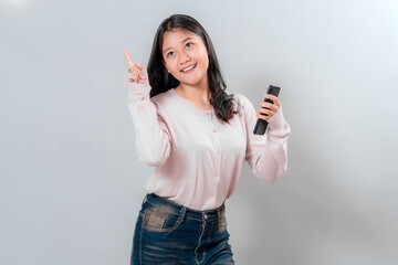 Young Asian woman smiling brightly while holding a smartphone and pointing up with confidence. Her...