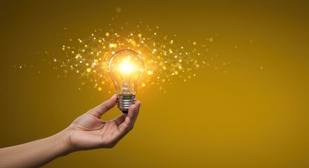Hand holding glowing lightbulb with sparkling particles on yellow background.
