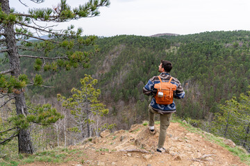 Young hiker with backpack standing on edge of cliff enjoying stunning vista of mountain forests on cloudy day