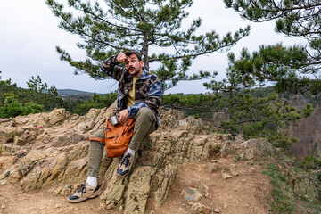 Tired hiker taking a break on a rocky mountain top, wiping sweat from his forehead under a pine tree