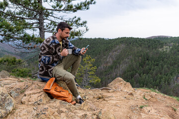 Naklejka premium Male hiker sitting on a rock, drinking coffee and using his phone while enjoying the mountain view