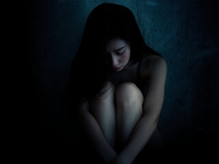 Obraz premium A young woman sits alone in a dark, shadowy corner with her arms wrapped around her knees, reflecting deep emotional pain, loneliness, and vulnerability in silence.
