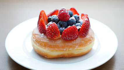 A glossy glazed donut sits on a white plate, adorned with vibrant strawberries, blueberries, and raspberries. Sunlight gently illuminates the scene, creating a warm atmosphere in a cafe