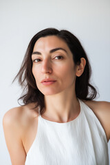 Fototapeta premium Portrait of a Confident 30-40 Year Old Woman with Dark Hair and Brown Eyes, Wearing a White Halter Neck Top Against a Clean White Background, Exuding Strength, Beauty and Self-Assuredness.