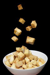 Tofu floats in the air and falls into a white bowl on a black background.