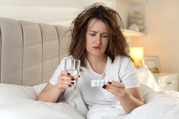 Unhappy woman with pills and glass of water suffering from hangover on bed at home