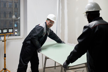 Two biracial professional construction workers putting huge plasterboard down carefully while...