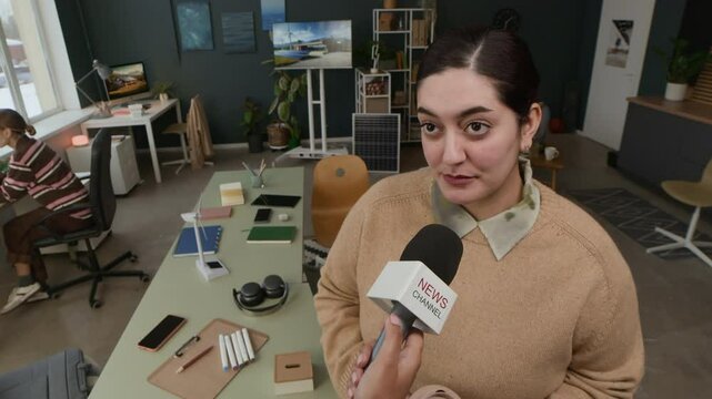 Female renewable energy engineer speaking into microphone to news channel reporter during interview in office, with models of solar panels and wind turbines in background