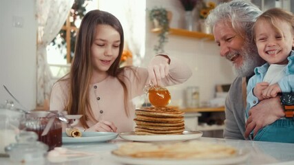 Joyful grandfather and his grandchildren at the table enjoy eating pancakes in the kitchen, cute girl pours honey over pancakes while little boy and grandpa laugh and have fun. - Powered by Adobe