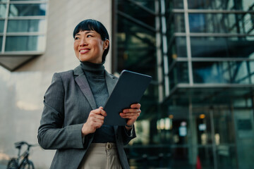 Smiling businesswoman holding tablet and looking away outside office building