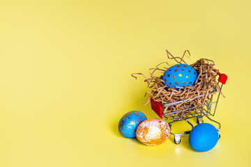 Easter eggs blue, gold, polka dot, blue, space colors, lies in a nest of paper in a shopping basket mini trolley, on a yellow background, beautiful Easter composition for Easter