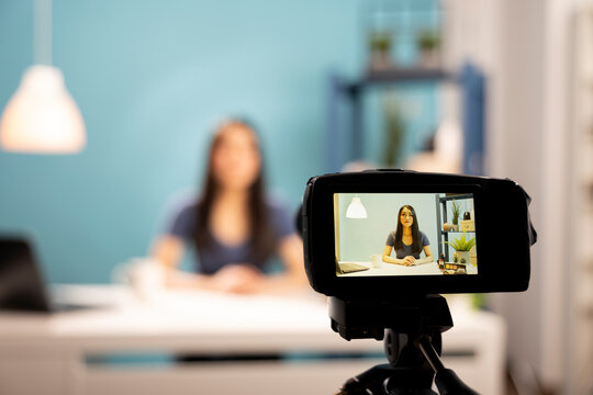 Selective focus of a camera on tripod recording a beauty vlogger seated at desk with blue background. This portrays behind the scenes of young woman, creating tutorial videos for subscribers.
