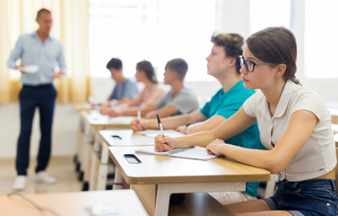 Portrait of focused teenage schoolgirl in glasses writing lectures in workbook in classroom during lesson
