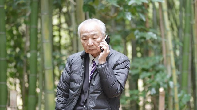 A winter park in Japan. A forest with lots of bamboo. A 78-year-old Japanese man in a suit is standing there. He is calling on his smartphone.