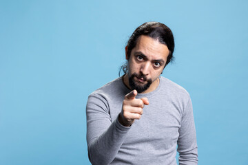 Male person with a serious attitude pointing at someone on camera, showing his index finger and choosing something or somebody against blue background. Young serious adult picks people.