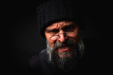 Stylish portrait of a confident mature man with his arms crossed in front of him, wearing a knitted hat and glasses. Staring, gray beard and black vest. Stylish discreet portrait on a dark background,