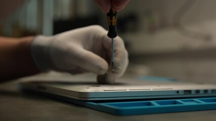 Technician wearing gloves using screwdriver to disassemble laptop computer on workbench, performing maintenance and repair tasks, close-up. Concept of computer hardware, repairing, upgrade, technology