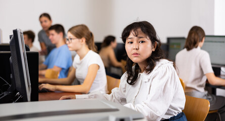 Young asian woman sitting in university computer room at workplace and looking at camera