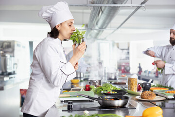 Dedicated female chef smelling the aroma of herbs and greenery, using tasty fresh rosemary or parsley leaves and sprinkling in the pan. Culinary expert cooking dishes with raw materials, gastronomy.