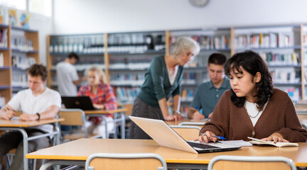 Obraz premium Asian fifteen-year-old schoolgirl, preparing for classes in the school library, studies on a laptop and writes notes in a copybook