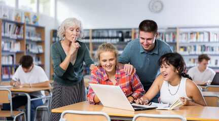 Adult librarian holding his finger on lips making silent gesture in library to laughing teenage pupils watching online tutorial video