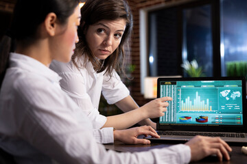 Two female colleagues collaborating on business analytics, reviewing data and discussing strategies on a laptop. They analyze financial information and plan tasks ahead in a professional workspace.