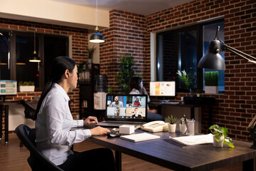 Asian female manager engages in a virtual meeting with her team, sharing business strategy ideas during a videoconference. The laptop shows her connecting with remote employees during her night shift.