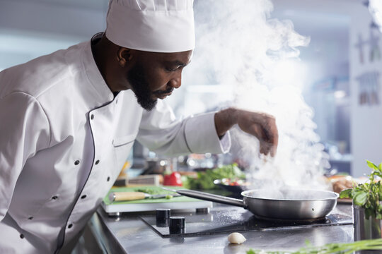 In a kitchen, african american chef adding the fresh herbs leaves to a plated dish. Male expert uses raw vegetables, spices and essential kitchen equipment to create a tasty meal.
