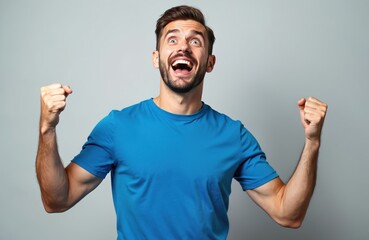Fototapeta premium Happy caucasian man celebrates success with raised arms clenched fists. Smiling face, excited expression, looking up with open mouth. Wearing blue t-shirt. Concept of win, triumph, joy, victory,
