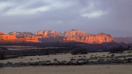 Panoramic view of the sunset light glowing on the cliffs of West Temple Mt. in Zion NP UT USA while the fields of Smith Mesa are in shadow and clouds almost fill the sky. 