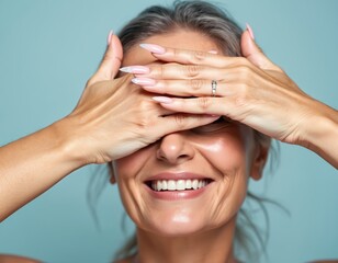 Mature woman covers eyes with manicured hands. Happy mature model with perfect skin and makeup. Portrait for beauty salon, aesthetic cosmetics, wellness, spa. Smiling lady in studio.