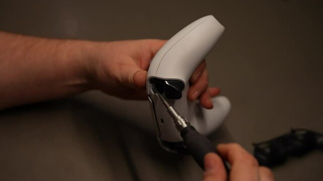 Close-up cropped shot of technician using screwdriver to disassemble and repair white game controller on gray table, demonstrating precision and concentration in electronics maintenance, slow motion.