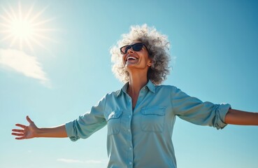 Happy senior woman with open arms smiles in sunlight. Elderly lady with grey hair, sunglasses enjoys sunny weather outdoors. Active lifestyle, positive mindset concept, happy aging, mental health.