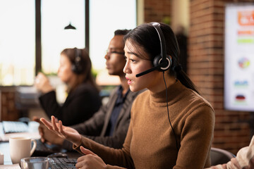 Asian businesswoman wearing wired headphones and providing assistance to remote clients. Serious female call operator with headset sitting at desk and handling customer inquiries.