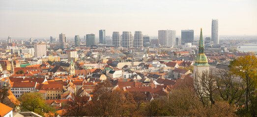 In historic district of Bratislava, Slovakia, tower of St. Martin Cathedral standing above red-tiled roofs, with Danube River and modern buildings visible in background on autumn day.