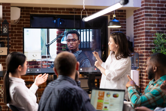 Diverse startup team working late in brick wall office, actively engaging in hybrid meeting with black businessman. Caucasian manager presents while others listen, with video call on digital screen.