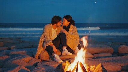 Romantic couple cuddling by a bonfire on the beach at night.