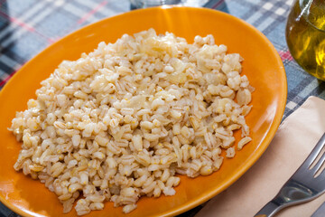 Boiled pearl barley served on yellow ceramic dish on checkered fabric surface..