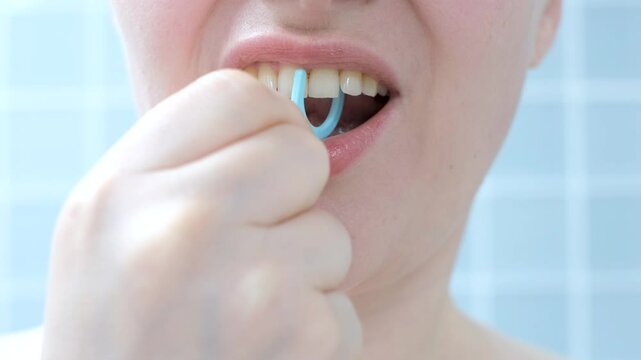 Young woman brushing teeth with disposable dental floss close-up against the background of blue tiles in the bathroom.