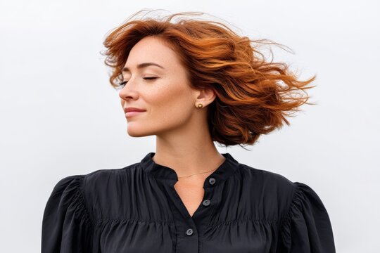 Redheaded woman in black shirt turning head with hair in motion and eyes closed in side view against sky blue background expressing emotional liberation serenity and individuality