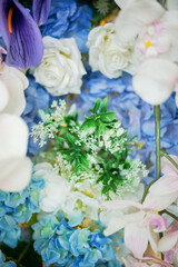 A vibrant close-up of a mixed floral arrangement featuring white roses, blue hydrangeas, purple Anthurium, and delicate white blossoms with green foliage. Suggests a festive or decorative display.