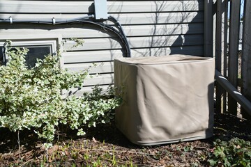 An outdoor central air conditioning condenser unit is covered with a protective beige storage cover outside a residential house.